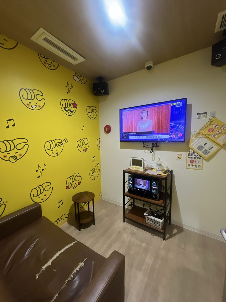 chocoZAPのセルフカラオケルーム内観。黄色い壁紙にキャラクターのイラスト、JOYSOUNDの機材とマイクが設置されている