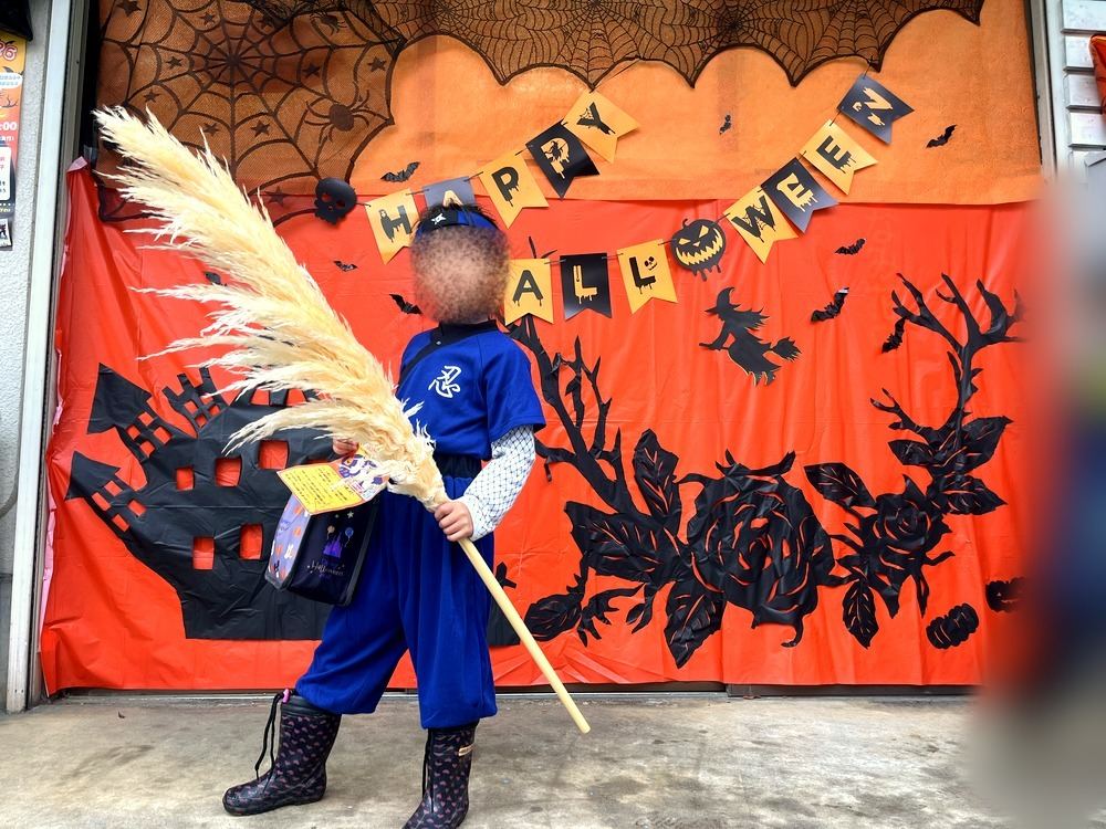 HAPPY HALLOWEENの飾りの前で、ススキのほうきを持ってポーズをとるニンジャの仮装の子ども
