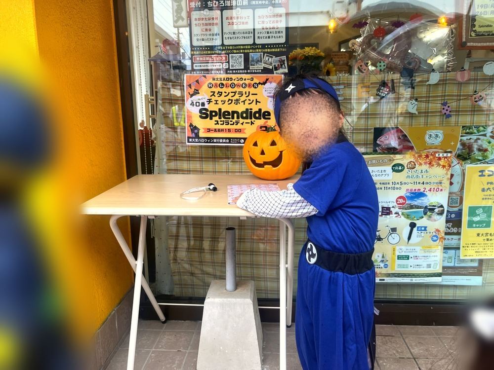 お店の前に置かれたスタンプラリーのチェックポイントの机とかぼちゃの飾り、ニンジャの仮装の子ども