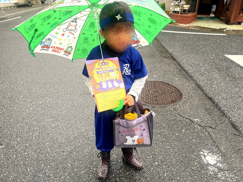雨の中、傘をさしてニンジャの仮装で東大宮ハロウィンウォークのカードを持つ子ども