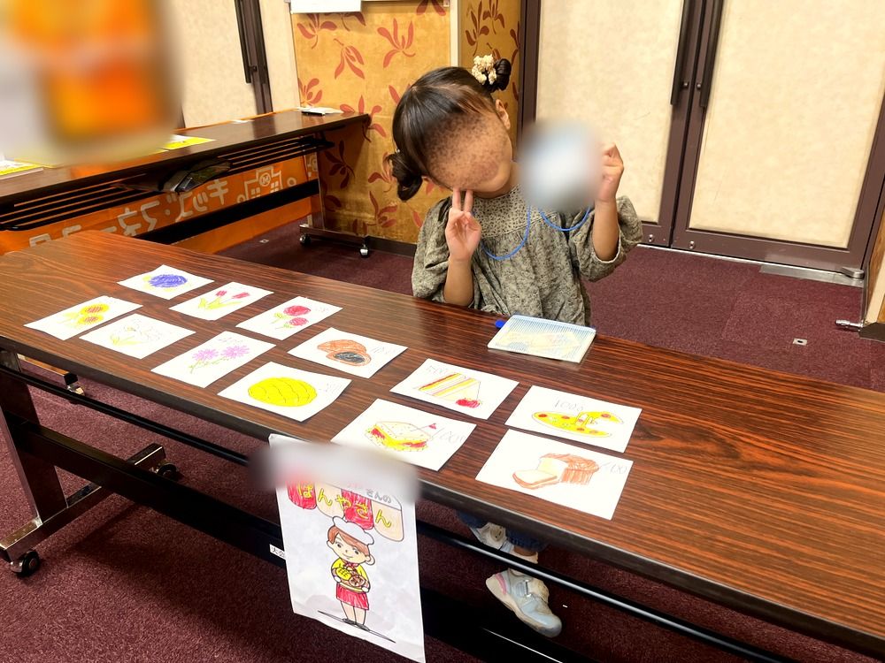 手描きの商品を並べた自分のパン屋さんの前でピースする5歳の子ども(顔はぼかし)