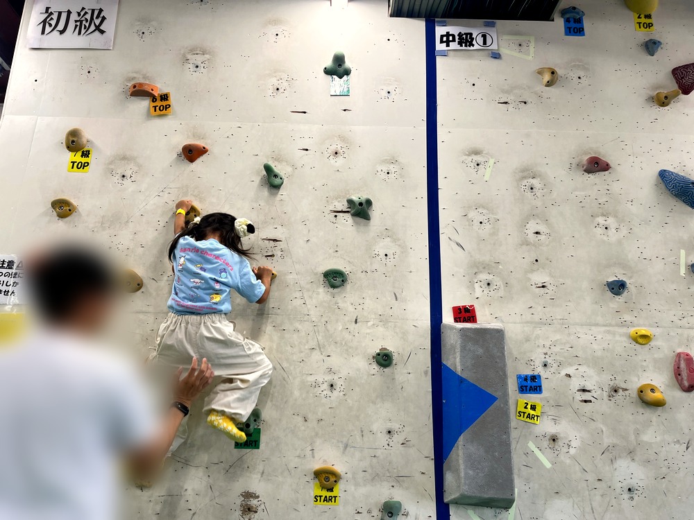 ボルダリングの壁を登る5歳の子どもと、下で支える大人